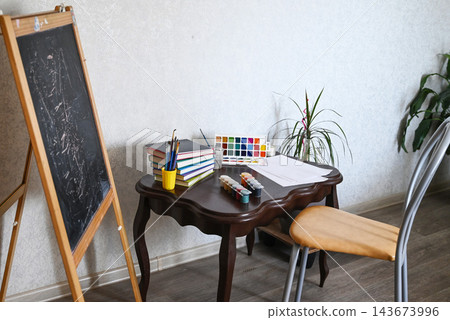 Art space includes a chair, chalkboard, easel, and plant in a room with plain walls and wooden floor Art space includes a chair, chalkboard, easel, and plant in a room with plain walls and wooden floor 143673996