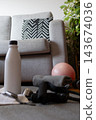 Home fitness items on a gray carpet floor including a water bottle, a small pink exercise ball, resistance bands, and a gray towel, ready for a workout near a couch and green plant 143674036