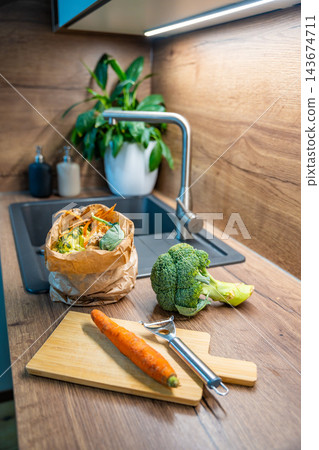 A paper bag of food scraps stands beside broccoli and a carrot at a kitchen sink. Separate biowaste collection supports composting in an urban apartment. A paper bag of food scraps stands beside broccoli and a carrot at a kitchen sink. Separate biowaste collection supports composting in an urban apartment. 143674711