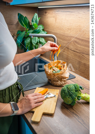 A woman holds carrot peelings above a paper bag of food scraps near a sink. Collecting vegetable waste supports composting in an urban apartment. A woman holds carrot peelings above a paper bag of food scraps near a sink. Collecting vegetable waste supports composting in an urban apartment. 143674713