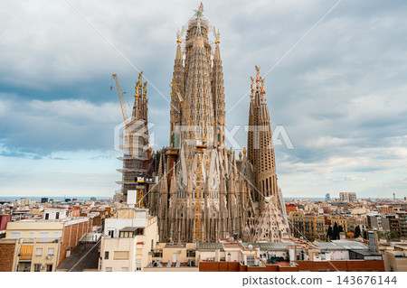 Sagrada Familia towers with construction cranes and scaffolding above Barcelona rooftops and the sea Sagrada Familia towers with construction cranes and scaffolding above Barcelona rooftops and the sea 143676144