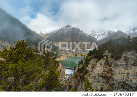 Aerial view of mountain valley with green lake, dam and sanctuary beneath snowy peaks in Vall de Nuria Aerial view of mountain valley with green lake, dam and sanctuary beneath snowy peaks in Vall de Nuria 143676317
