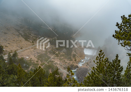 Mountain lake dam spilling water into a rocky gorge in fog seen from above in Vall de Nuria Mountain lake dam spilling water into a rocky gorge in fog seen from above in Vall de Nuria 143676318