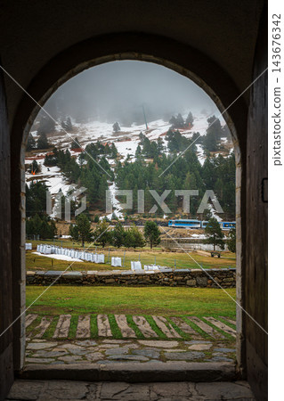 Blue mountain train passing pine trees below a snow patched ski slope seen through a stone arch in Vall de Nuria Blue mountain train passing pine trees below a snow patched ski slope seen through a stone arch in Vall de Nuria 143676342