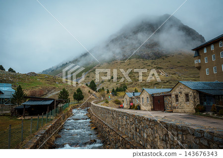 Mountain stream flowing between stone walls past old stone buildings below a fog covered peak in Vall de Nuria Mountain stream flowing between stone walls past old stone buildings below a fog covered peak in Vall de Nuria 143676343