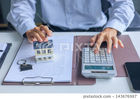 Businessman using calculator and touching miniature house with contract document on the table. Concept for real estate investment, loan, rent, mortgage, insurance Businessman using calculator and touching miniature house with contract document on the table. Concept for real estate investment, loan, rent, mortgage, insurance 143676987