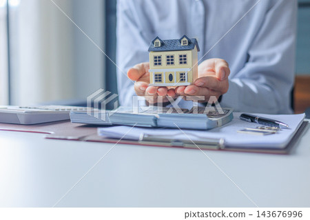 Businessman, Banker, Realtor at office desk holding a house model with money, calculator, and financial documents or contract on the table Businessman, Banker, Realtor at office desk holding a house model with money, calculator, and financial documents or contract on the table 143676996