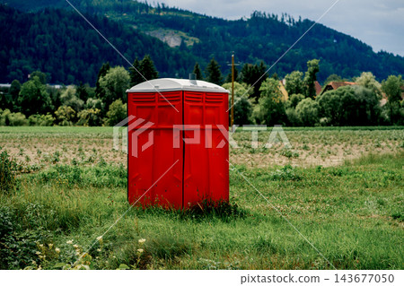 A red portable toilet stands in a grassy field with crops and mountains behind. Concept of sanitation, outdoor facilities, worksite and convenience. A red portable toilet stands in a grassy field with crops and mountains behind. Concept of sanitation, outdoor facilities, worksite and convenience. 143677050