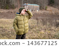 A preteen boy in a green jacket shields his eyes with his hand and gazes into the distance, standing in a field outside the city with an autumn forest in the background. 143677125
