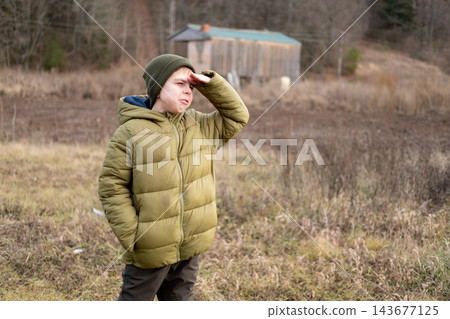 A preteen boy in a green jacket shields his eyes with his hand and gazes into the distance, standing in a field outside the city with an autumn forest in the background. A preteen boy in a green jacket shields his eyes with his hand and gazes into the distance, standing in a field outside the city with an autumn forest in the background. 143677125