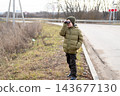 A schoolboy in a green jacket looks through binoculars while standing by a paved country road in autumn. 143677130