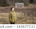 A portrait of a smiling preteen boy in a green jacket, standing in a field in front of an old wooden barn in the autumn countryside 143677131