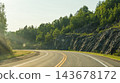 Curving paved road with double yellow lines beside rocky forested hillside in warm summer sunlight, scenic rural roadway landscape 143678172
