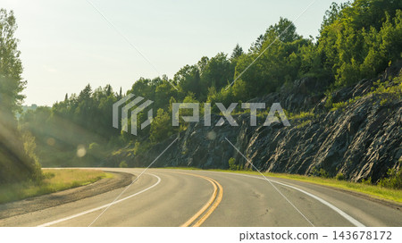 Curving paved road with double yellow lines beside rocky forested hillside in warm summer sunlight, scenic rural roadway landscape 143678172