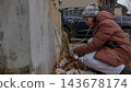 Woman in brown puffer jacket kneeling beside a concrete wall, removing weeds from a snowy garden with a parked car visible in the background 143678174