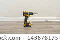 Yellow and black cordless power drill standing upright on light wood flooring beside a blank wall in an unfurnished interior 143678175