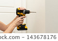 Person holding a yellow and black cordless drill against a white wall beside an interior door in a home improvement scene 143678176