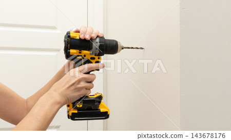 Person holding a yellow and black cordless drill against a white wall beside an interior door in a home improvement scene Person holding a yellow and black cordless drill against a white wall beside an interior door in a home improvement scene 143678176