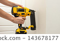 Person holding a yellow cordless nail gun with attached magazine against a white paneled door in a close up tool demonstration 143678177