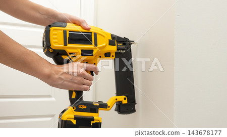 Person holding a yellow cordless nail gun with attached magazine against a white paneled door in a close up tool demonstration 143678177