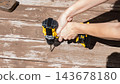 Hands holding a cordless drill above weathered wooden planks with scattered tools in a bright outdoor repair workspace 143678180