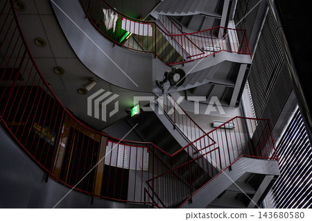 這是一張建築物結構的照片,拍攝角度是從建築物內部的樓梯井向上仰望,可以看到紅色的欄桿和灰色的鋼製樓梯層層疊疊。 這是一張建築物結構的照片,拍攝角度是從建築物內部的樓梯井向上仰望,可以看到紅色的欄桿和灰色的鋼製樓梯層層疊疊。 143680580
