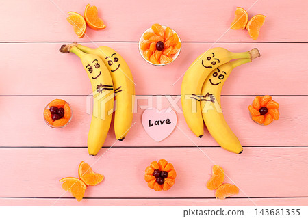 Adorable bananas in love, hugging each other, accompanied by a small heart labeled love and surrounded by butterflies and flowers made from mandarin oranges. This playful treat is perfect for children Adorable bananas in love, hugging each other, accompanied by a small heart labeled love and surrounded by butterflies and flowers made from mandarin oranges. This playful treat is perfect for children 143681355