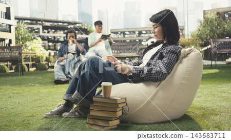 Student, reading and books as a woman relaxes on a beanbag with an open book and stacked textbooks nearby on a lawn. Independent learning, literacy and campus life suit peaceful study outdoors. 143683711