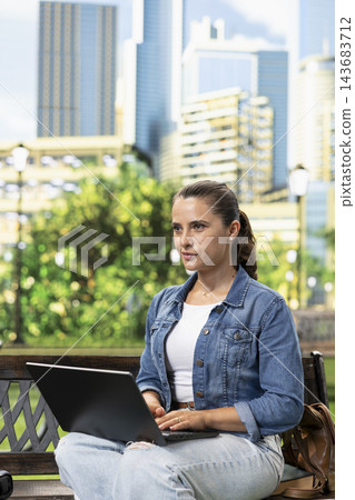 Woman, laptop and remote work as casually dressed professional types on computer while seated on a wooden bench. Productivity, flexibility and mobile working suit urban park surroundings. Woman, laptop and remote work as casually dressed professional types on computer while seated on a wooden bench. Productivity, flexibility and mobile working suit urban park surroundings. 143683712