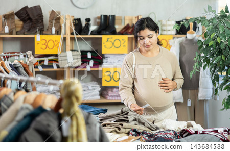 Pregnant mulatto young female client choosing trousers in clothing mall 143684588