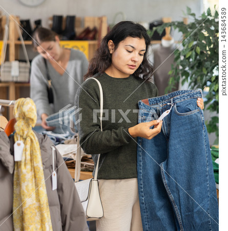 Happy latina female client carefully choosing stylish jeans trousers in clothing store. Shopping and sales concept Happy latina female client carefully choosing stylish jeans trousers in clothing store. Shopping and sales concept 143684589