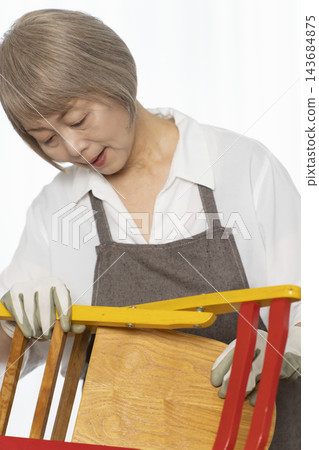 A senior woman remakes a doll chair in her living room as a DIY hobby. A senior woman remakes a doll chair in her living room as a DIY hobby. 143684875