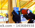 Electrician worker drills into an electrical box mounted on wooden post beneath covered work area during electrical repair 143684938