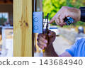 An electrician connects wires in switch box mounted on wooden post at an outdoor site 143684940