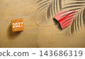 Red paper airplane flying over sand background with number 2027 on wooden cube and palm tree shadow. Concept of Happy New Year 143686319