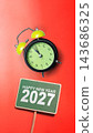 Happy New Year 2027 message on a green sign with a black and lime green alarm clock pointing almost to midnight, set against a solid bright red background. Concept of Happy New Year 143686325