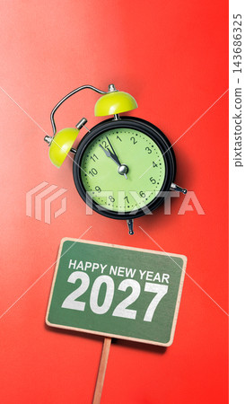 Happy New Year 2027 message on a green sign with a black and lime green alarm clock pointing almost to midnight, set against a solid bright red background. Concept of Happy New Year 143686325