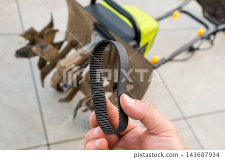 A metal tiller blade rests on a patio surface, while a person holds a replacement rubber belt designed for garden machinery. Stone tiles, indicating an outdoor work area. A metal tiller blade rests on a patio surface, while a person holds a replacement rubber belt designed for garden machinery. Stone tiles, indicating an outdoor work area. 143687934