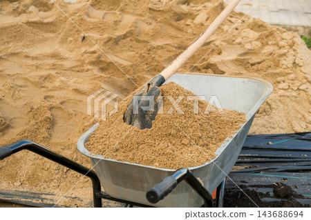 A wheelbarrow is prominently positioned at a construction site, loaded with sandy soil and featuring a shovel placed on top. 143688694