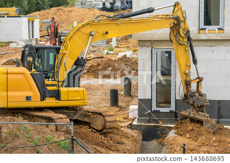 The foundation of a building is positioned near the site where soil is being excavated. Heavy machinery is used to prepare the earth for ongoing construction work. The foundation of a building is positioned near the site where soil is being excavated. Heavy machinery is used to prepare the earth for ongoing construction work. 143688695