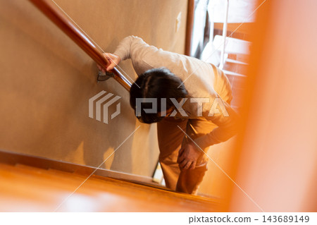A middle-aged/senior woman with mobility issues climbs the stairs at home, holding onto the handrail. A middle-aged/senior woman with mobility issues climbs the stairs at home, holding onto the handrail. 143689149