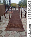 Rusted metal accessibility ramp with brown railings leads up to a building entrance in autumn. Background features colorful fall trees. The ramp surface is expanded metal grating. 143691003