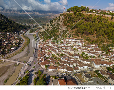 Berat Albania spreads along Osum River beneath wooded hills, with white Ottoman era houses clustered below historic castle walls. Aerial view highlights red tile roofs and mountain valley. Berat Albania spreads along Osum River beneath wooded hills, with white Ottoman era houses clustered below historic castle walls. Aerial view highlights red tile roofs and mountain valley. 143691085