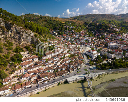 Berat Albania spreads across hillside beneath bright clouds, with traditional white houses and red tiled roofs. Historic townscape follows riverfront road beside green hills and stone riverbanks. 143691086