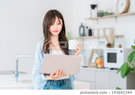 一位年輕女子一邊看著廚房裡的筆記型電腦，一邊手捧咖啡，臉上露出微笑。輕鬆愉快的日常生活。 143691344
