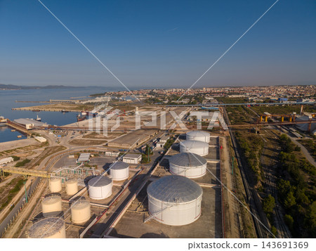 Coastal oil storage terminal stands beside harbor, with large fuel tanks, pipelines and rail sidings across industrial grounds. Port facilities and city buildings line blue waterfront under clear sky. Coastal oil storage terminal stands beside harbor, with large fuel tanks, pipelines and rail sidings across industrial grounds. Port facilities and city buildings line blue waterfront under clear sky. 143691369