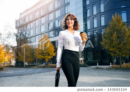 Attractive woman carrying coffee and business bag with confidence. Pretty woman wearing elegant office clothing near modern business center. Executive lifestyle concept. 143693104