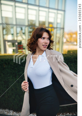 Beautiful woman adjusting elegant blazer with confident business appearance. Attractive woman standing near modern office building outdoors. Corporate fashion concept. 143693143