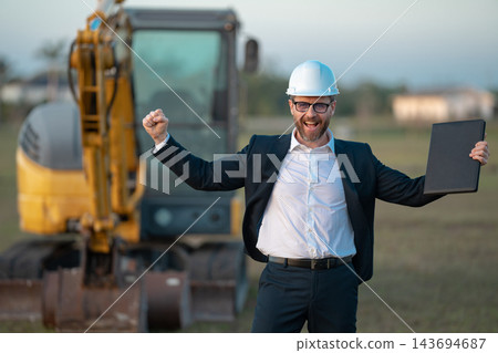 Construction business owner. Man in suit and hardhat halmet at building construction site. Civil engineer worker in front of house near excavator. Architect, supervisor investor, construction manager. 143694687
