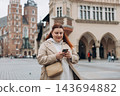 Attractive young female tourist is exploring new city. Redhead girl holding a smart phone on Market Square in Krakow. Traveling Europe in summer. St. Marys Basilica. The Cloth Hall, Vacation concept 143694882